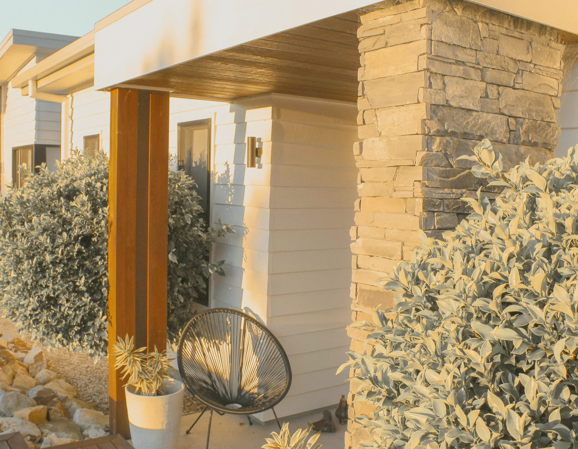 The front porch of a new house in low sun, silver leaved shrubs and a potted succulent among river stones