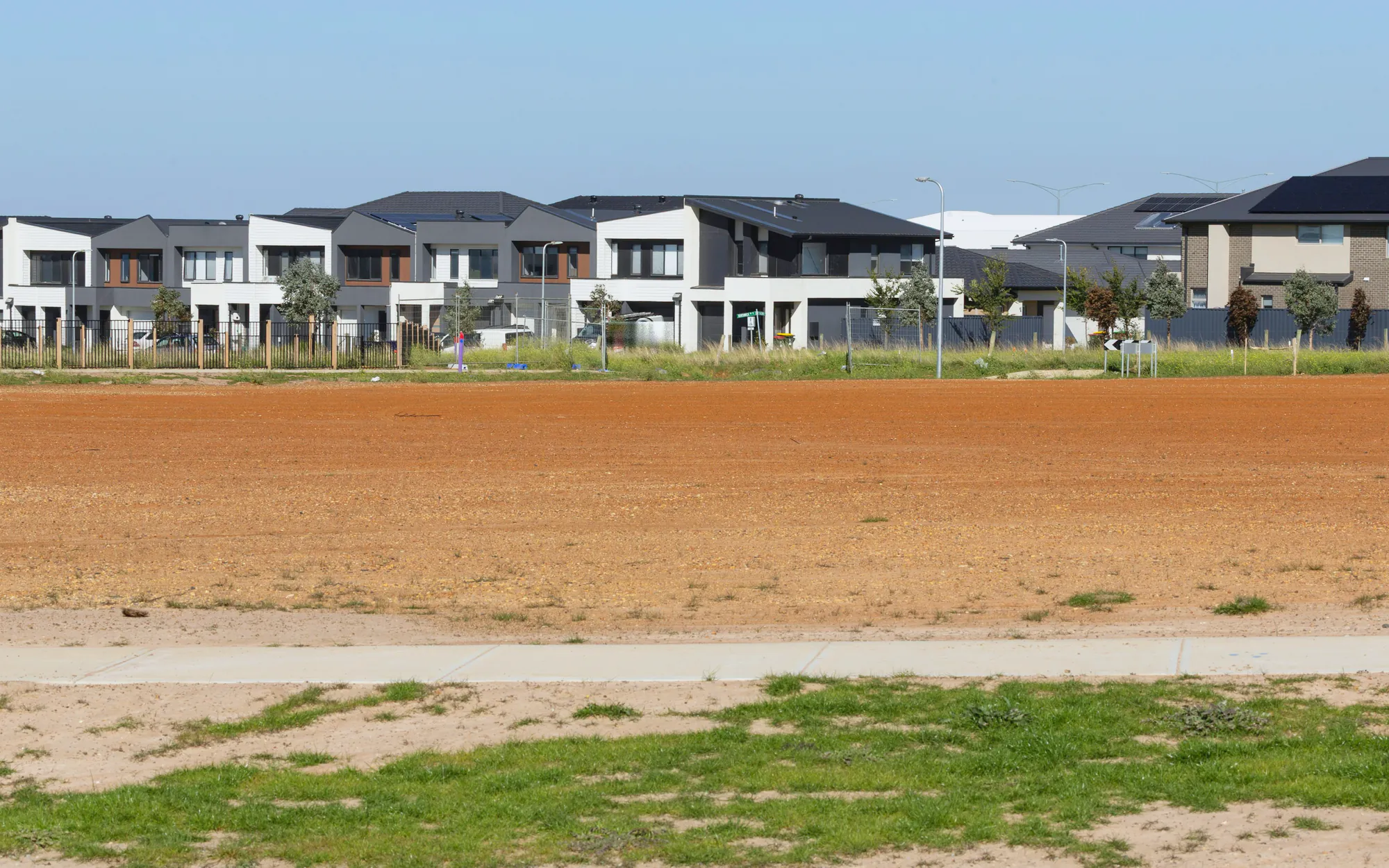 A levelled bare block of red earth under a clear sky, new two storey houses along its far side: a garden before it is begun